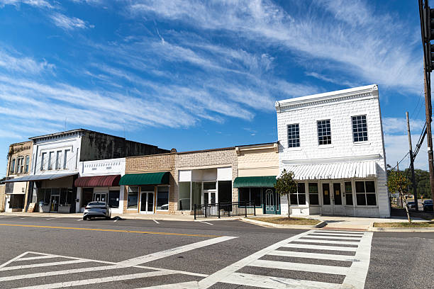 A row of historic commercial buildings lines the main street in downtown Eutaw, Alabama, featuring classic early 20th century architecture with brick and painted facades. The buildings include storefronts with green awnings, large windows, and traditional small-town commercial design elements. Eutaw, the county seat of Greene County, is known for its well-preserved historic downtown district and antebellum architecture. A crosswalk and street parking are visible in the foreground under partly cloudy skies during the day.