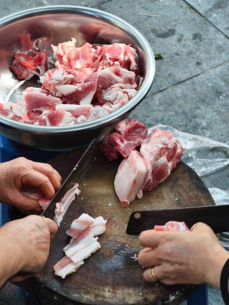 Hands cutting raw pork meat on a wooden chopping board, with more meat in a silver bowl
