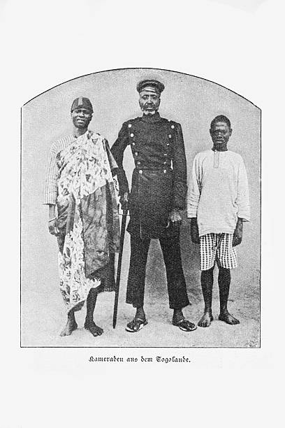 Authentic black and white photograph from the late 19th century depicting three men from German Togoland. The German caption reads 'Kameraden aus dem Togolande' (Comrades from Togoland). The central figure wears a colonial-era uniform, while the men flanking him wear traditional and everyday clothing. A studio portrait from the German colonial period in Africa.