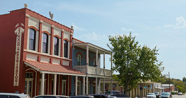 Historic downtown Brenham, Texas features well-preserved 19th-century commercial buildings along the main street. The red brick Victorian-era structure with ornate white trim displays classic architectural details including arched windows and decorative cornices. A blue building with white columns and wraparound balcony represents typical Texas commercial architecture from the late 1800s. Tree-lined streets and period lampposts complement the historic district atmosphere in this Washington County seat, known for its German heritage and antique shopping district.