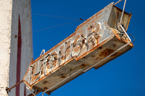 A weathered vintage neon sign displays faded lettering against a clear blue sky in downtown Brenham, Texas. The metal sign shows significant rust and aging, with glass neon tubing still visible despite years of exposure to the elements. Located in Washington County, Brenham is known as the home of Blue Bell Creameries and features a historic downtown district with many preserved buildings from the late 19th and early 20th centuries. The town's commercial district reflects its heritage as an important railroad and agricultural center in the Brazos Valley region.