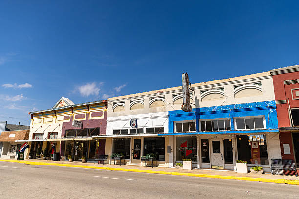 A row of historic commercial buildings lines Main Street in downtown Brenham, Texas, displaying the characteristic architecture of a small Texas town's central business district. The colorful storefronts feature a mix of late 19th and early 20th century facades with arched windows, decorative cornices, and varied paint schemes including burgundy, white, and bright blue. The buildings house various local businesses and shops typical of a historic downtown area. Brenham, known as the home of Blue Bell Creameries, maintains its small-town charm through preservation of its historic downtown core. Clear blue skies with scattered white clouds provide a backdrop to this well-preserved example of Texas Main Street architecture.