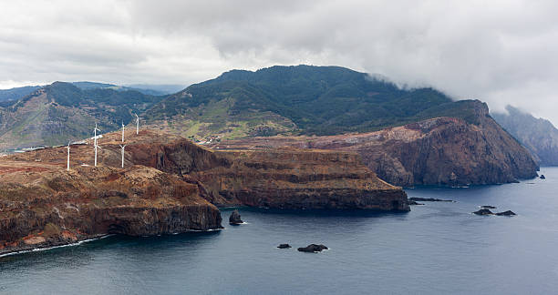 Wind turbines on Madeira's rugged coastline. Atlantic Ocean waves crash against rocky cliffs. Mountainous landscape with green hills under cloudy sky.