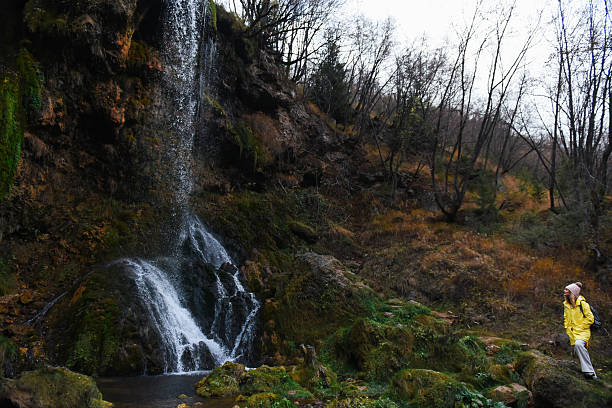 Woman wearing a yellow raincoat and backpack hikes beside a mossy waterfall in an autumn forest, pausing on a rocky trail to admire the scenic, peaceful landscape during a cool day.