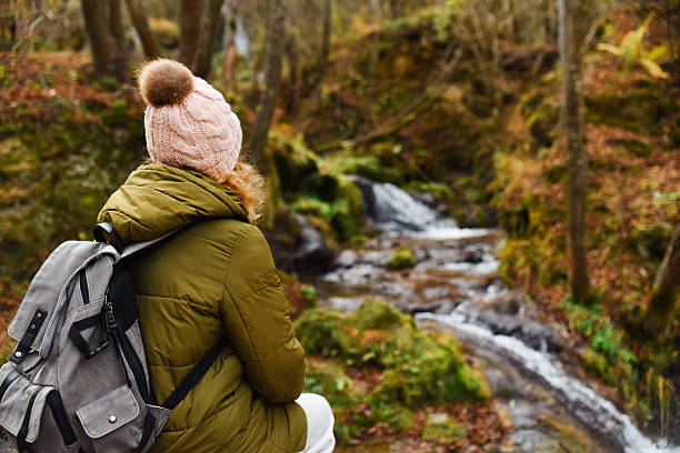 A woman hiker in a green puffer jacket and pink beanie sits with her backpack beside a mossy forest stream, enjoying a peaceful autumn walk and quiet outdoor moment of reflection.