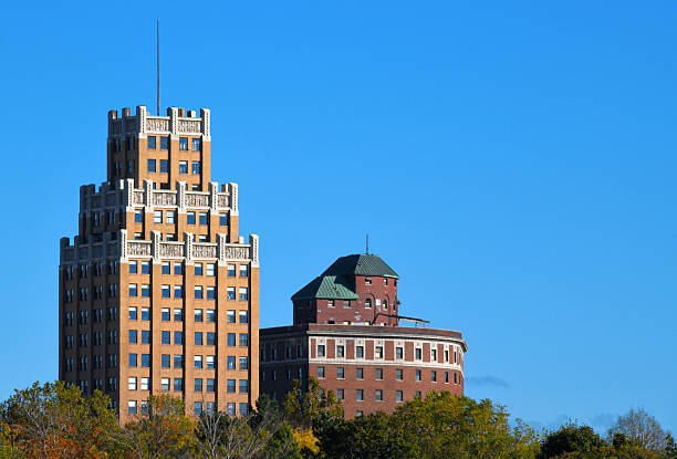Niagara Falls - New York State, USA: on the left is the former United Office Building, an Art Deco skyscraper completed in 1929, characterized by its tan brick facade, stepped setbacks, and ornate terracotta detailing. It currently operates as The Giacomo, a boutique hotel and residential building. On the right stands the Hotel Niagara, a twelve-story commercial structure opened in 1924, distinguished by its red brick exterior and a rounded, multi-story bay section topped with a green roof. The foreground features the tops of various trees displaying early autumn foliage in shades of green and yellow.