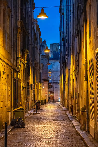 Warm-lit narrow Bordeaux street at dusk featuring cobblestone pavement, hanging lamps, historic stone façades and a quiet European alleyway atmosphere during blue hour evening light.