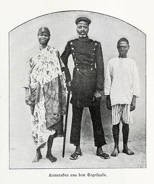 Vintage black and white photograph depicting three men from Togoland, West Africa, posing for a portrait during the German colonial era, circa late 19th or early 20th century. The central figure, an older man with a goatee, wears a dark, double-breasted uniform and a peaked cap. To his left, a man is dressed in traditional West African attire, including a patterned cloth. To his right stands a younger man in a simple shirt and checkered shorts. The original German caption reads 'Kameraden aus dem Togolande,' which translates to 'Comrades from Togoland.'