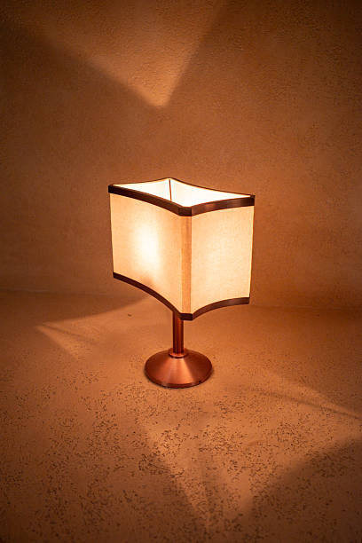 Warm light casts soft shadows from a square lamp. Minimalist design features cream-colored fabric shade. Metal base and stem offer sleek, modern contrast