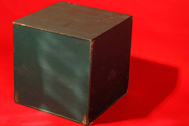 Dark brown cube with worn edges sits on a bright red background. The cube casts a soft shadow to the right side. Its surface shows texture and uneven coloration