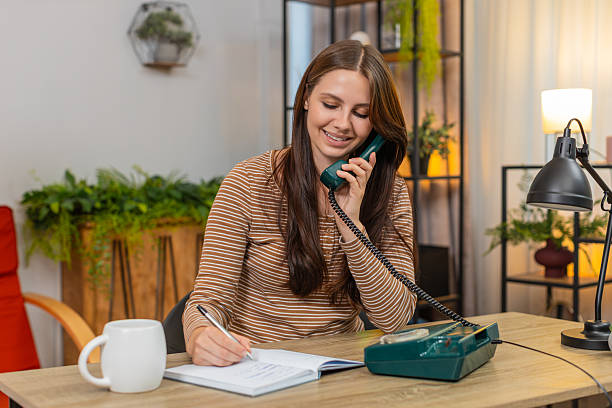 Young woman at home calls on retro landline phone from 90s nods listens and writes address in notebook. Caucasian girl at desk says thank you notes details with pen confirms time clearly and ends call