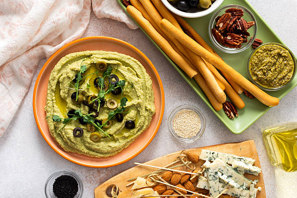 Broccoli hummus is artistically displayed in a bowl, garnished with olives. Accompanied by a charcuterie board featuring cheese, nuts, and breadsticks for a delightful snack experience