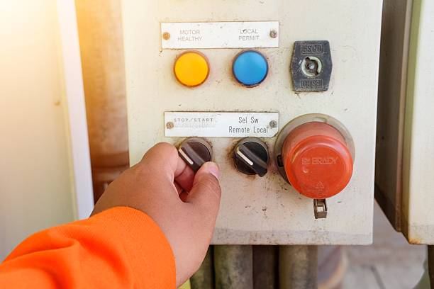 Close-up of a hand in an orange sleeve adjusting a switch on a vintage industrial control panel with warning lights and emergency stop.