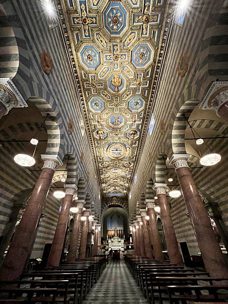 Volterra Cathedral's nave extends towards the altar, showcasing a coffered ceiling with intricate blue and gold details, marble columns, and patterned walls creating a historical atmosphere