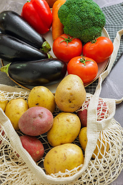 Variety of fresh farm produce with eggplant, red bell pepper, broccoli, carrots, ripe tomatoes, and mixed potatoes in a reusable bag