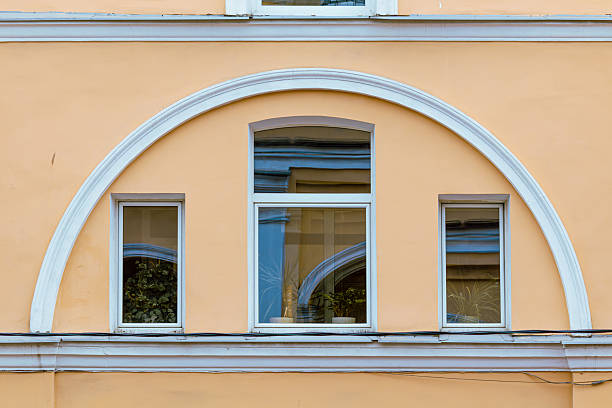 Three aligned windows with a shared white arch create a balanced architectural composition. Warm wall color, reflections, and indoor plants add charm and visual depth to the scene.