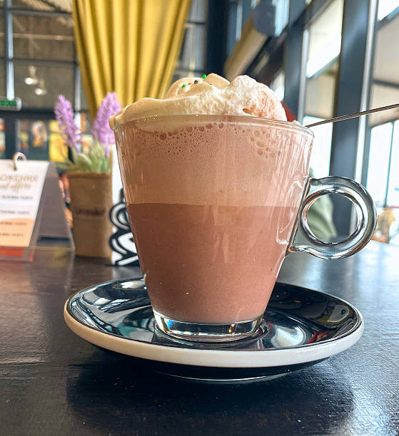 Hot chocolate topped with whipped cream and colorful sprinkles served in a clear glass cup on a black and white saucer, with a cozy cafe background