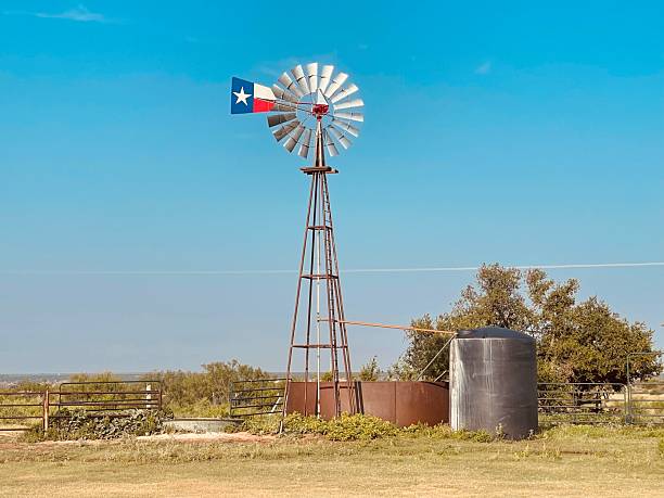 A traditional windmill stands in a quiet rural landscape beneath a clear blue sky, accented by a Texas flag, symbolizing independence, heritage, and life across the open American countryside.