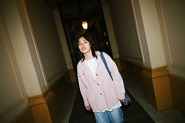 Asian person standing in a quiet indoor building corridor at dusk, wearing casual pink shirt and blue jeans with a shoulder bag