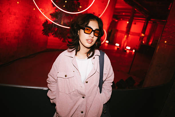 Young woman wearing sunglasses and a pink shirt looking at the camera in a dark room with red neon lighting