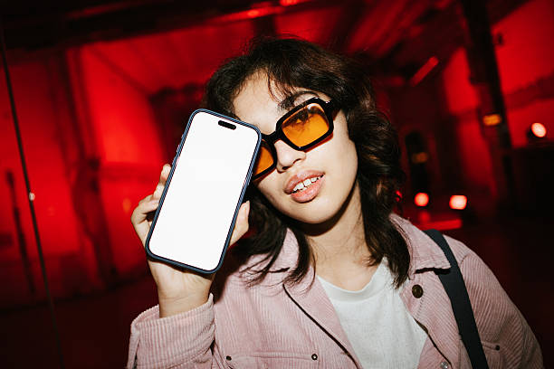 Young woman wearing trendy sunglasses and a stylish jacket holding a modern smartphone with a blank screen, ideal for mockups