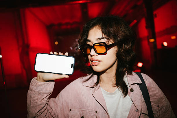 Young woman in stylish sunglasses holding a smartphone with a blank white screen up against a vibrant red lit background