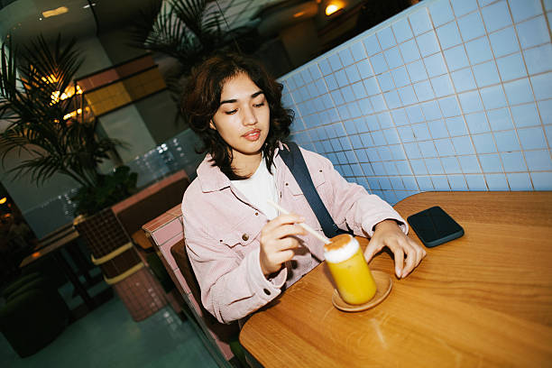 Young woman in pink jacket sipping a refreshing cold beverage with a straw at a wooden table in a stylish cafe