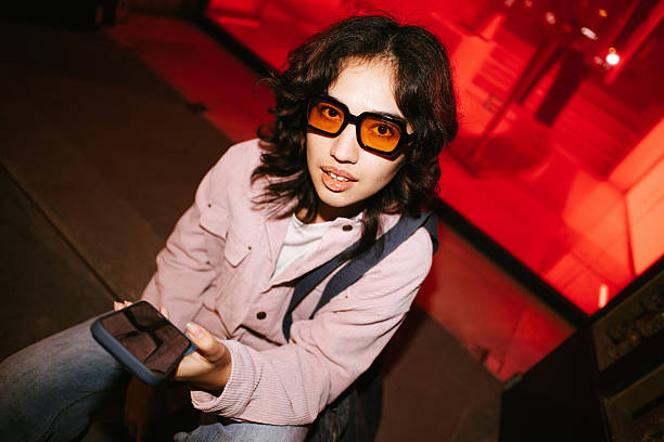Young woman in pink jacket and orange retro sunglasses holding a smartphone, looking at the camera in urban red light