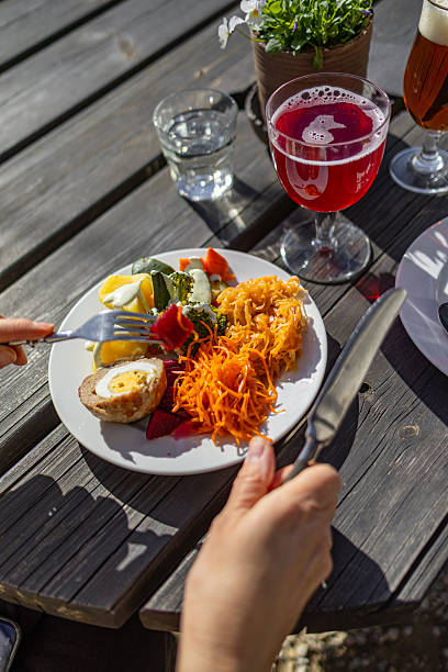 Bright outdoor meal with hands holding cutlery over a plate of vegetables, egg dish, and shredded carrots, paired with a vivid red drink on a rustic wooden table. Natural light and slight copy space
