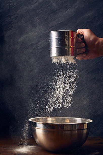 Man's hand sifting wheat flour through a sieve mug at table on black background, close-up