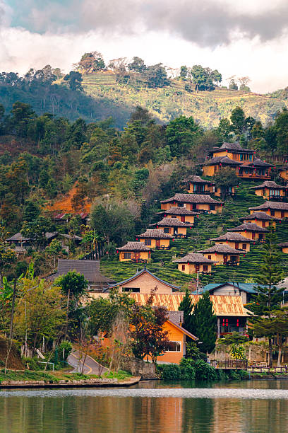 A scenic view of a Chinese-style accommodation on a hillside amidst tea plantations in Ban Rak Thai, Mae Hong Son, Thailand. A large reservoir stands in the foreground, its reflection visible in the water. There is a wooden pavilion in the middle of the water and a walkway along the reservoir.