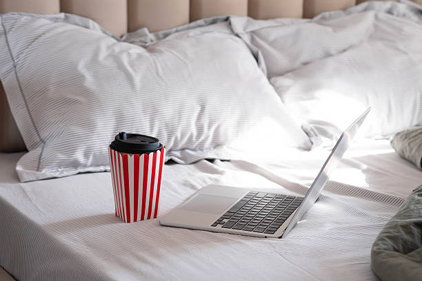 A laptop on a neatly made bed with striped pillows. Next to it a red-and-white striped coffee cup. Soft daylight creates a cozy work-from-home atmosphere.