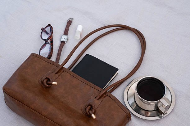 Brown leather tote bag with a black notebook inside, paired with tortoiseshell glasses, leather watch, lip balm and a metal cup of coffee on a light fabric background. Minimalist still life.