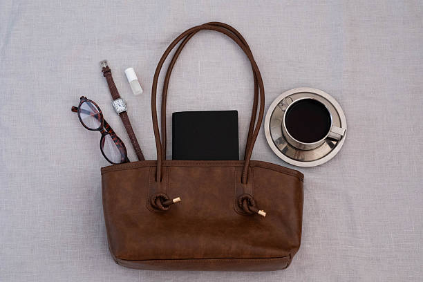 Brown leather tote bag with a black notebook inside, paired with tortoiseshell glasses, leather watch, lip balm and a metal cup of coffee on a light fabric background. Minimalist still life.
