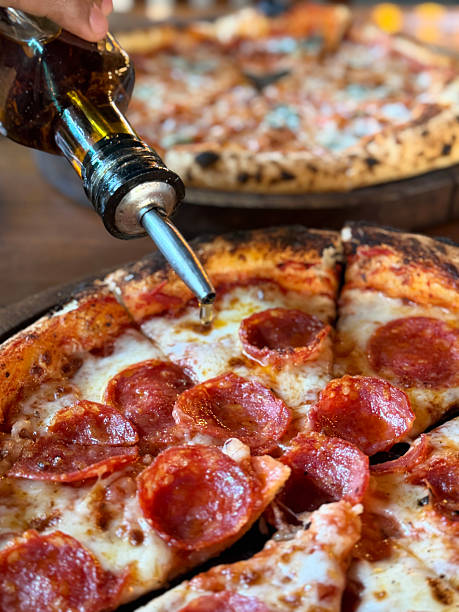 Close up image of drip from metal olive oil pourer spout onto sliced pepperoni pizza on wooden pizza paddle, melted golden buffalo mozzarella cheese, rich tomato marinara sauce and spicy salami topping and crispy crust, elevated view, focus on foreground
