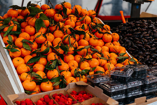 Fresh mandarins with green stems displayed in a market box alongside organic berries and figs. The image represents healthy eating, organic produce, and local market shopping.