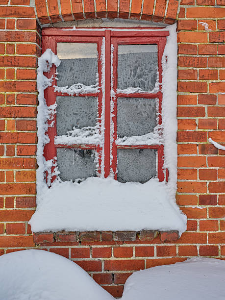 A red window frame rests in a brick wall, dusted with fresh snow along the sill and frost on the glass panes. The scene exudes a quiet, cold winter atmosphere and rustic charm, hinting at old Danish architecture and enduring texture.