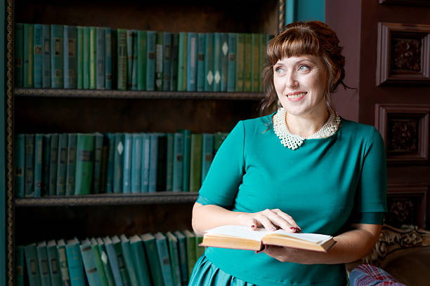 A happy adult woman in a green dress and pearl collar holding an open book and looking away with a smile. Classic library background with rows of books. Concept of inspiration and lifelong learning.
