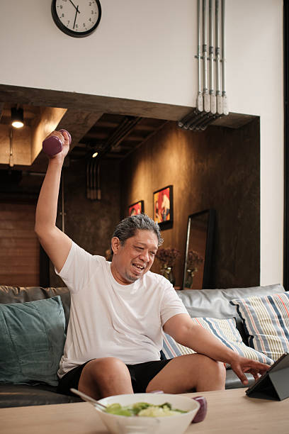Fat Asian mature man raises dumbbells, watching healthy social media channel.