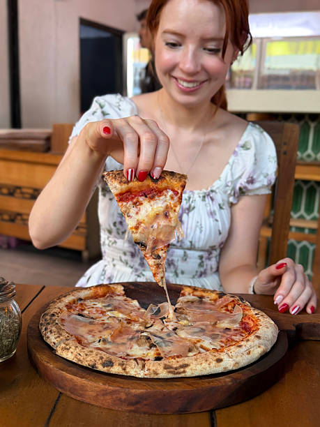 Image attractive, redheaded woman holding thin, crispy crust Prosciutto Crudo pizza, slice raised from wooden pizza paddle, melted golden buffalo mozzarella cheese, rich tomato sauce, displaying manicure nail art, restaurant setting, focus on foreground