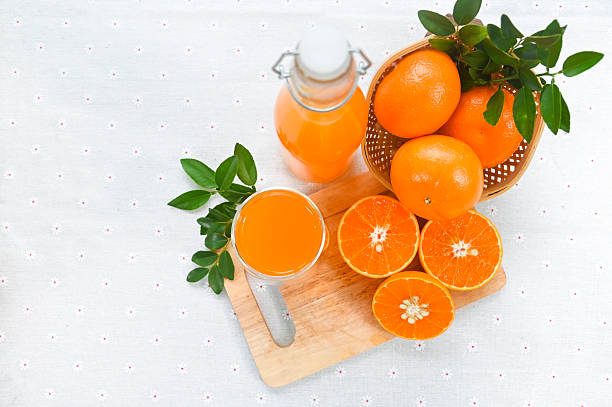 An overhead view captures a glass of freshly squeezed orange juice, a bottle of fresh juice, and whole and halved oranges arranged on a white background.