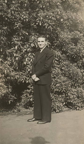 Black and white vintage portrait of a young man wearing a suit standing in front of dense leafy foliage in the 1930s.