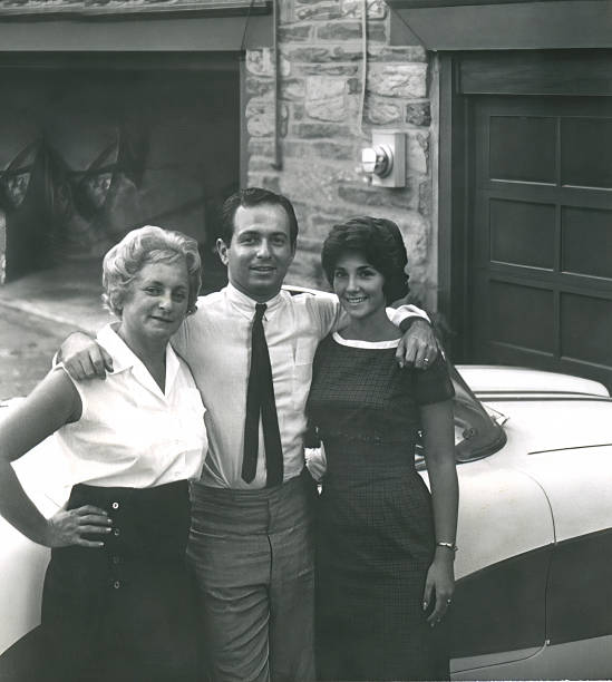 Black and white vintage photo of two women and a man posing together beside a classic automobile outside a home around 1960.