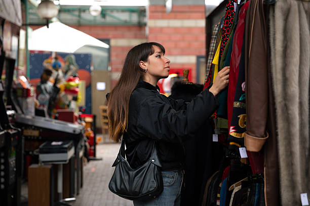 Young woman browsing clothing on a rack at a flea market in Paris, France. The candid lifestyle scene shows a shopper carefully examining garments at a second-hand clothing stall inside an urban market. The image highlights vintage fashion culture, sustainable shopping, and the everyday atmosphere of Paris flea markets where visitors explore unique clothing and retro styles.