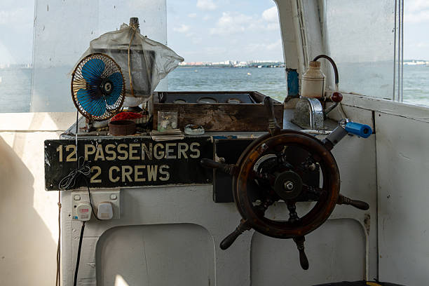Wooden helm rests beside weathered control panel. Black sign denotes capacity: twelve passengers, two crew. Fan, tools, and ropes adorn cluttered vessel interior. Window frames distant water and sky