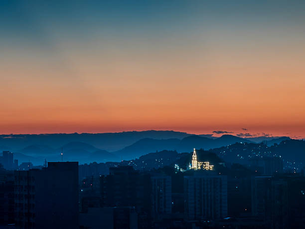 Illuminated Saint Antony Lisboa Church at dawn in Rio de Janeiro. A city skyline at dusk with buildings in the foreground and mountains in the background. The sky is illuminated with a warm orange hue, and the buildings are silhouetted against the horizon.