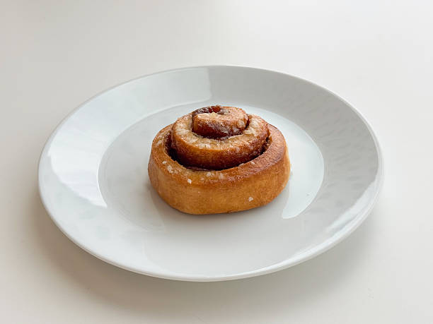 Cinnamon roll pastry dusted with sugar served on white plate against minimal background. Bakery product styling, sweet breakfast idea, cafe menu item, dessert simplicity, clean food presentation concept