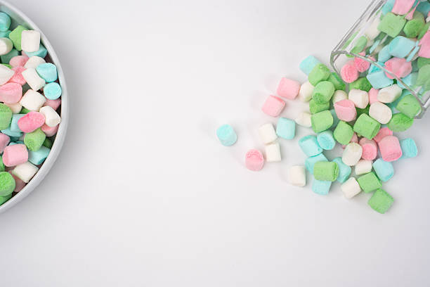 Small pink, green, blue, and white marshmallows are presented in a light gray bowl. Additional candies scatter from a glass onto a bright white background, ready for a sweet snack