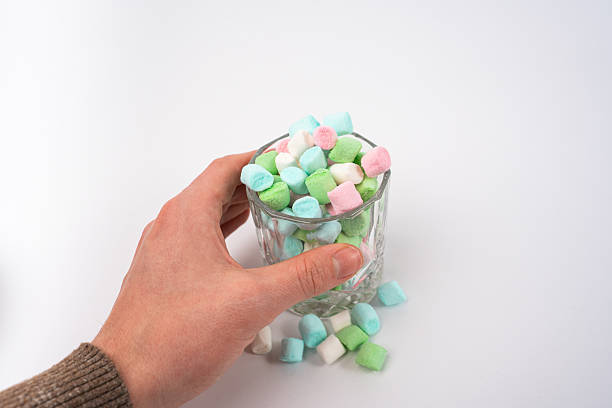 A hand holds a clear glass overflowing with small, soft pastel colored marshmallows. Several sweet candies in pink, green, and blue have spilled onto the smooth white surface below