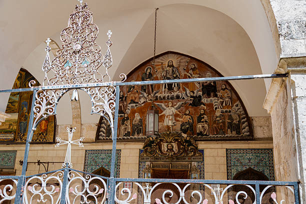 Elaborate painted religious murals depicting Christ, apostles, and angels adorn an arched facade at a historic Christian church in the Old City of Jerusalem. Intricate white wrought iron gates topped with decorative crosses and scrollwork stand in the foreground, while glazed ceramic tilework and baroque gilded ornamentation frame the entrance below. Ideal for editorial and commercial use relating to religion, pilgrimage, Middle Eastern heritage, and sacred architecture.